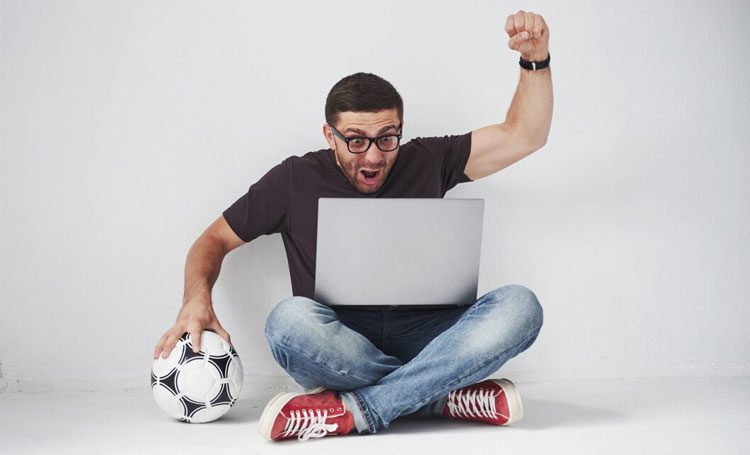 Imagem de homem sentado com notebook no colo comemorando vitória em aposta esportiva, com bola de futebol na mão
